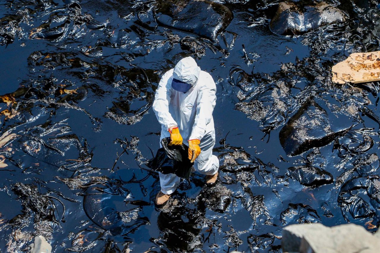 Trabajador con traje blanco y guantes amarillos sobre rocas cubiertas de petróleo
